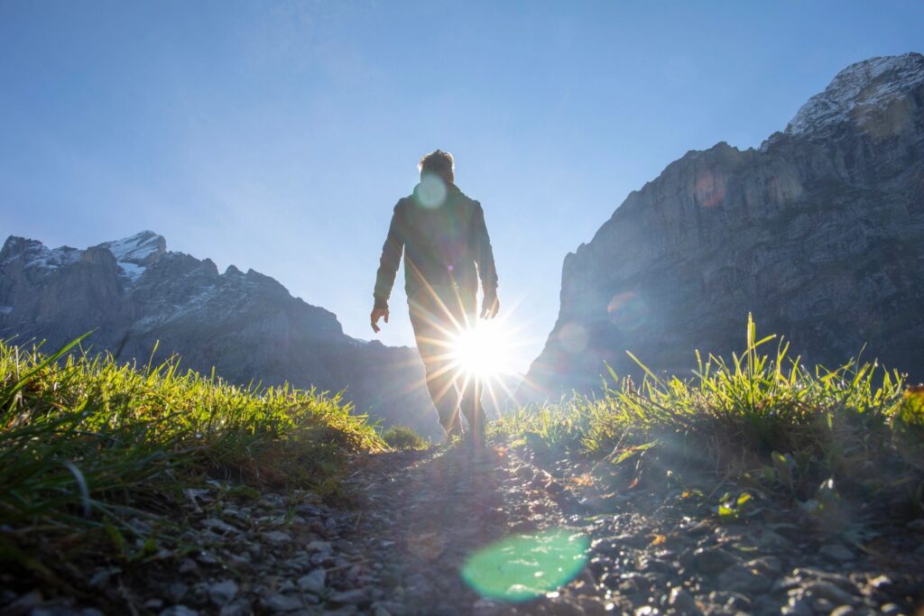 A person walking a mountain path towards the low sun, light spilling across the grass